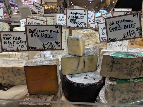 Various types of cheese are displayed on a counter in a market or store. Each cheese has a label with its name, origin, and price. The cheeses vary in color and texture, and the signs indicate different types of milk sources and flavor profiles.