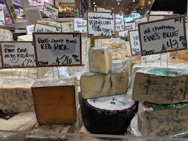 Various types of cheese are displayed on a counter in a market or store. Each cheese has a label with its name, origin, and price. The cheeses vary in color and texture, and the signs indicate different types of milk sources and flavor profiles.