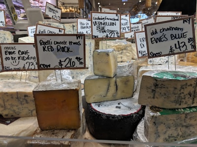 Various types of cheese are displayed on a counter in a market or store. Each cheese has a label with its name, origin, and price. The cheeses vary in color and texture, and the signs indicate different types of milk sources and flavor profiles.