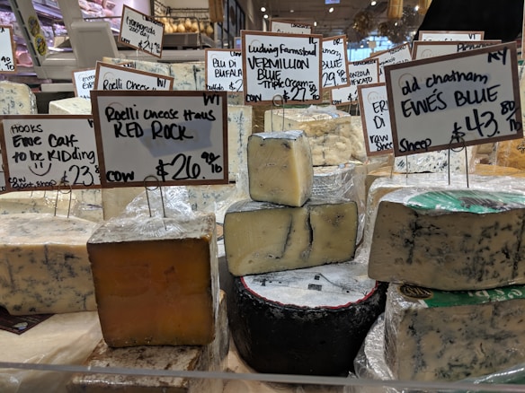 Various types of cheese are displayed on a counter in a market or store. Each cheese has a label with its name, origin, and price. The cheeses vary in color and texture, and the signs indicate different types of milk sources and flavor profiles.