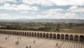 An expansive view of a wide stone courtyard with a series of arches along one side. Several people are walking or standing on the courtyard, enjoying the panoramic view of a lush, rural landscape with fields, trees, and distant mountains under a partly cloudy sky.