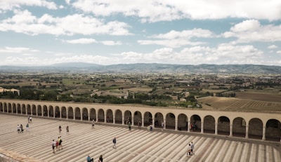 An expansive view of a wide stone courtyard with a series of arches along one side. Several people are walking or standing on the courtyard, enjoying the panoramic view of a lush, rural landscape with fields, trees, and distant mountains under a partly cloudy sky.