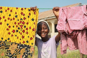 A child wearing a headband and a light purple shirt smiles brightly while holding up colorful fabrics on a clothesline outside. The fabric on the left has a vibrant yellow pattern with red and black stars, and the fabric on the right is a red and white checkered pattern. The background features a building with a slanted roof.