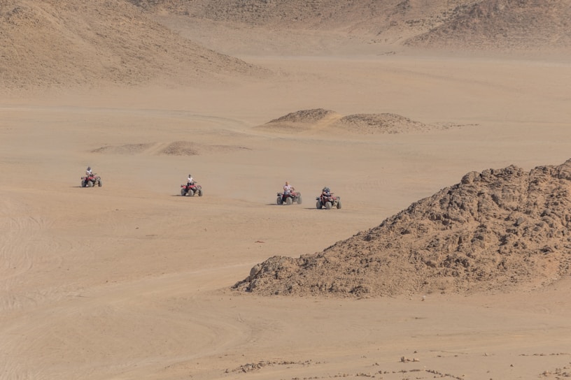 Four individuals riding all-terrain vehicles traverse a vast, arid landscape with sandy terrain and rocky outcrops. The environment appears to be desert-like, with light brown hues dominating the scene.