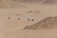 Four individuals riding all-terrain vehicles traverse a vast, arid landscape with sandy terrain and rocky outcrops. The environment appears to be desert-like, with light brown hues dominating the scene.