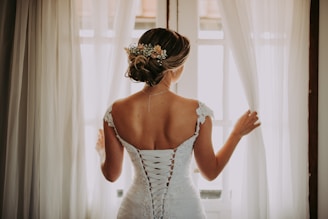 woman in wedding dress standing near window looking outside while holding the curtains