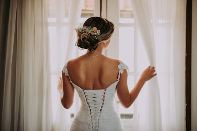 woman in wedding dress standing near window looking outside while holding the curtains