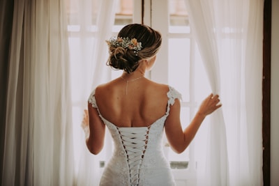 woman in wedding dress standing near window looking outside while holding the curtains
