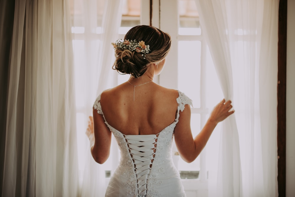 Frau im Hochzeitskleid steht in der Nähe des Fensters und schaut nach draußen, während sie die Vorhänge hält