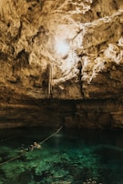 A natural cave with a pool of clear turquoise water at the bottom. Sunlight streams in from an opening above, illuminating the rugged, rocky walls. Two people wearing snorkel gear are swimming in the pool, with a rope extending across the water.