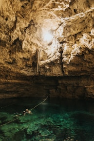 A natural cave with a pool of clear turquoise water at the bottom. Sunlight streams in from an opening above, illuminating the rugged, rocky walls. Two people wearing snorkel gear are swimming in the pool, with a rope extending across the water.