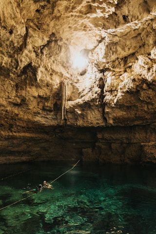 A natural cave with a pool of clear turquoise water at the bottom. Sunlight streams in from an opening above, illuminating the rugged, rocky walls. Two people wearing snorkel gear are swimming in the pool, with a rope extending across the water.