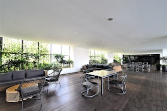 A spacious indoor lounge area with modern furniture arrangements, including sofas, armchairs, and tables. Large windows bring in natural light and reveal lush greenery outside. The room has wooden flooring and a white ceiling with recessed lighting.