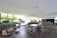 A spacious indoor lounge area with modern furniture arrangements, including sofas, armchairs, and tables. Large windows bring in natural light and reveal lush greenery outside. The room has wooden flooring and a white ceiling with recessed lighting.