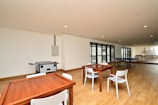 A spacious indoor recreational area with wooden flooring, featuring a foosball table, ping pong tables, and wooden tables and chairs. Large windows line one wall, allowing natural light to fill the space.
