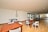 A spacious indoor recreational area with wooden flooring, featuring a foosball table, ping pong tables, and wooden tables and chairs. Large windows line one wall, allowing natural light to fill the space.
