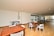 A spacious indoor recreational area with wooden flooring, featuring a foosball table, ping pong tables, and wooden tables and chairs. Large windows line one wall, allowing natural light to fill the space.
