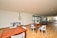 A spacious indoor recreational area with wooden flooring, featuring a foosball table, ping pong tables, and wooden tables and chairs. Large windows line one wall, allowing natural light to fill the space.
