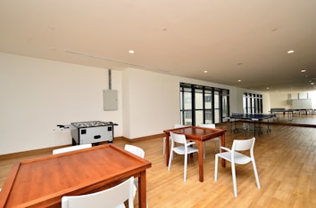 A spacious indoor recreational area with wooden flooring, featuring a foosball table, ping pong tables, and wooden tables and chairs. Large windows line one wall, allowing natural light to fill the space.
