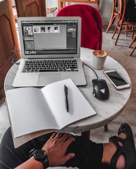 A cozy workspace setup on a round marble table featuring a laptop displaying image editing software. Beside the laptop is a wireless mouse, a smartphone, a notebook with a pen placed over it, and a cup of coffee. A red jacket is draped over a chair in the background. The person using the setup has a hand resting on their knee, wearing a wristwatch.