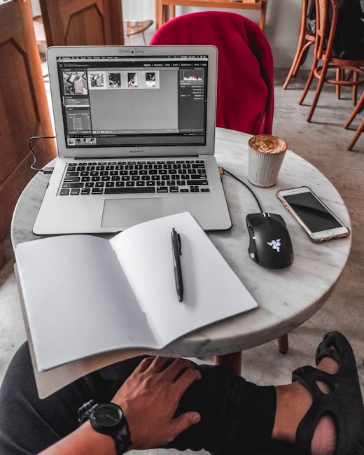 A cozy workspace setup on a round marble table featuring a laptop displaying image editing software. Beside the laptop is a wireless mouse, a smartphone, a notebook with a pen placed over it, and a cup of coffee. A red jacket is draped over a chair in the background. The person using the setup has a hand resting on their knee, wearing a wristwatch.