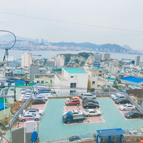 A view of a cityscape with a parking lot in the foreground, filled with cars and a small truck. The background features a densely packed urban area with various buildings, and a harbor with cranes and ships visible in the distance, set against a backdrop of hills.