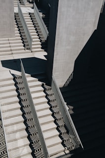 Concrete staircases with metal railings are illuminated by sunlight, casting geometric shadows along the steps. The perspective creates an abstract visual effect with sharp contrasts between light and dark areas.