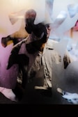 A person is partially visible behind a frosted glass or translucent surface, creating an abstract and mysterious effect. The lighting casts shadows and highlights, emphasizing the texture of the glass and the person's silhouette.