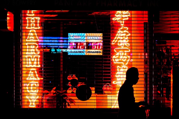 Bright neon signs in orange and yellow light up the storefront of a pharmacy, visible through metal security bars. The silhouette of a person walking by is prominent in the foreground, set against the vibrant colors. Additional signage displays a lottery number in bright white and blue.