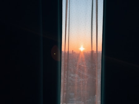 A sunrise view through a partially open window with sheer curtains, showing the skyline of a city with buildings silhouetted against an orange and blue sky.
