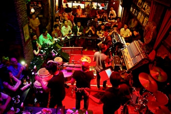 A lively music venue filled with people enjoying a live performance. The setting features a band with various musical instruments like drums, keyboards, and guitars on a stage with red lighting. The audience is seated closely around the stage, providing an intimate atmosphere. The room is dimly lit with warm, ambient lighting, enhancing the cozy and energetic vibe of the event.