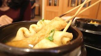 Skewered fish cakes are immersed in a savory broth within a dark bowl. The fish cakes have a golden-brown hue, garnished with green onions. In the background, a person is partially visible, wearing a dark red top, suggesting a dining setting.