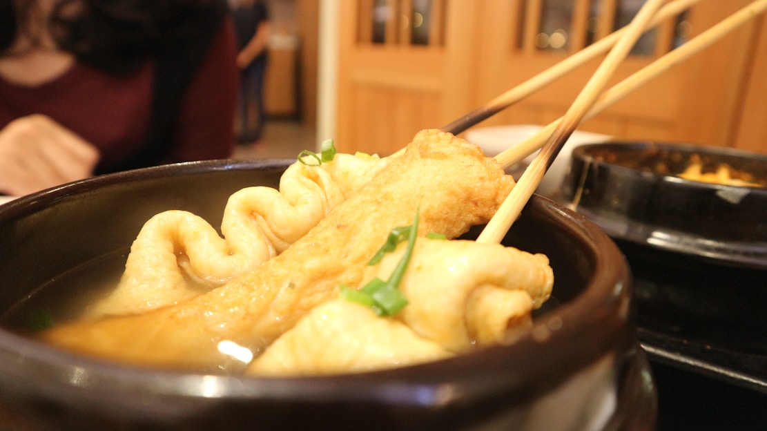Skewered fish cakes are immersed in a savory broth within a dark bowl. The fish cakes have a golden-brown hue, garnished with green onions. In the background, a person is partially visible, wearing a dark red top, suggesting a dining setting.