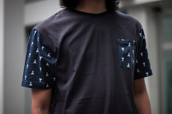 A person wearing a dark T-shirt with patterned sleeves and pocket stands, partially visible from the neck down. The material appears smooth, and there are small, repeated white figures on the sleeves and pocket.