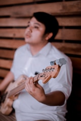 A person is holding a guitar with a focus on the instrument's neck and headstock, featuring a blurred wooden wall background. The individual is wearing a white shirt.