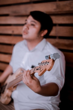 A person is holding a guitar with a focus on the instrument's neck and headstock, featuring a blurred wooden wall background. The individual is wearing a white shirt.