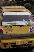 A yellow minibus with signs of wear and tear, displaying the numbers 19 and 238 on its front. The bus has a roof rack, large windshield, side mirrors, and a yellow license plate reading 'BK 1194 FY'. The background reveals some greenery and a building with a tiled roof.