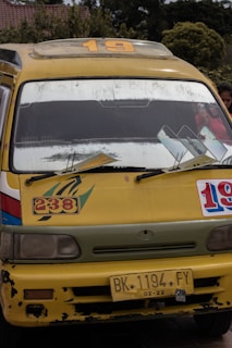 A yellow minibus with signs of wear and tear, displaying the numbers 19 and 238 on its front. The bus has a roof rack, large windshield, side mirrors, and a yellow license plate reading 'BK 1194 FY'. The background reveals some greenery and a building with a tiled roof.