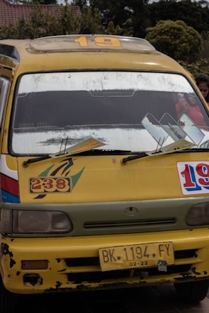 A yellow minibus with signs of wear and tear, displaying the numbers 19 and 238 on its front. The bus has a roof rack, large windshield, side mirrors, and a yellow license plate reading 'BK 1194 FY'. The background reveals some greenery and a building with a tiled roof.