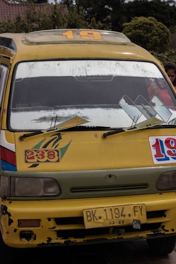 A yellow minibus with signs of wear and tear, displaying the numbers 19 and 238 on its front. The bus has a roof rack, large windshield, side mirrors, and a yellow license plate reading 'BK 1194 FY'. The background reveals some greenery and a building with a tiled roof.