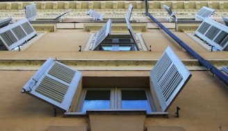 A low-angle view of a building facade with multiple windows featuring open white shutters. The windows are aligned vertically along a beige wall and are flanked by decorative architectural details, like cornices and moldings. A downspout runs along the side of the building.