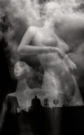 Two mannequins are positioned against a backdrop of swirling clouds, creating a surreal and dreamlike atmosphere. The scene blends elements of urban architecture silhouetted in the foreground with reflections of natural elements, blurring the lines between the artificial and the natural.
