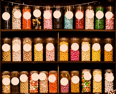 Rows of cylindrical glass jars filled with colorful candies are neatly arranged on shelves. The jars have white circular labels, and the candies inside the jars come in various colors and shapes. The image gives a visually appealing and organized display of sweet treats.