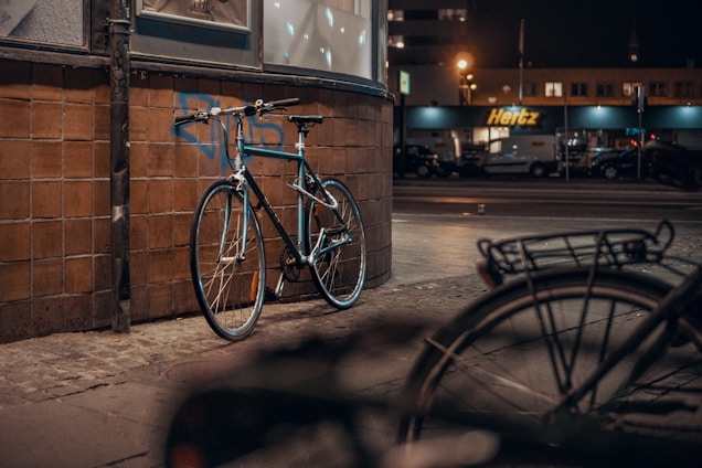 A bicycle is leaning against a tiled wall on a dimly lit street. In the background, there is a rental car service center with illuminated signage. The street appears empty, with a second bicycle blurred in the foreground.