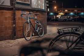 A bicycle is leaning against a tiled wall on a dimly lit street. In the background, there is a rental car service center with illuminated signage. The street appears empty, with a second bicycle blurred in the foreground.