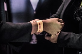 A close-up of two hands, one wearing a yellow wristband with red text. Both individuals are dressed in formal dark clothing.