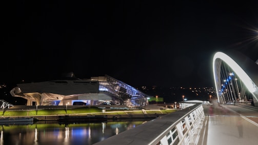 A modern bridge illuminated with bright lights spans across a water body at night. On the left, a futuristic building with angular architecture is also well-lit, showcasing a blend of glass and metal. The reflections of lights are visible in the water beneath.