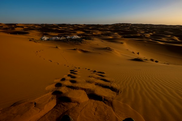 A vast expanse of sand dunes stretching into the horizon under a clear blue sky. Footprints are visible leading towards a small cluster of tents that appear to be set up as a camp. The sand looks golden in the sunlight, and the overall landscape is serene and unspoiled.