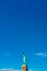 A monumental statue of a robed woman holding a torch in her right hand stands on a pedestal against a clear blue sky. The statue is green in color and detailed, symbolizing liberty.