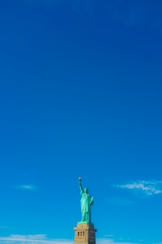 A monumental statue of a robed woman holding a torch in her right hand stands on a pedestal against a clear blue sky. The statue is green in color and detailed, symbolizing liberty.